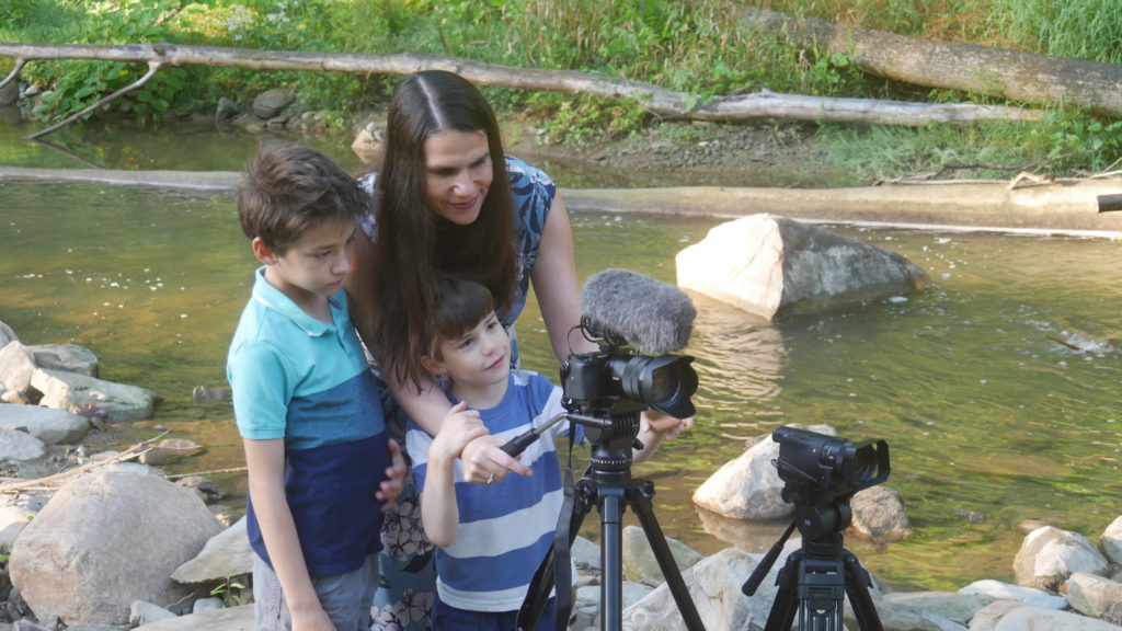 Alexandra and her two young sons are standing by a river, all crowded behind a camera with a mic. They are all wearing blue shirts. 