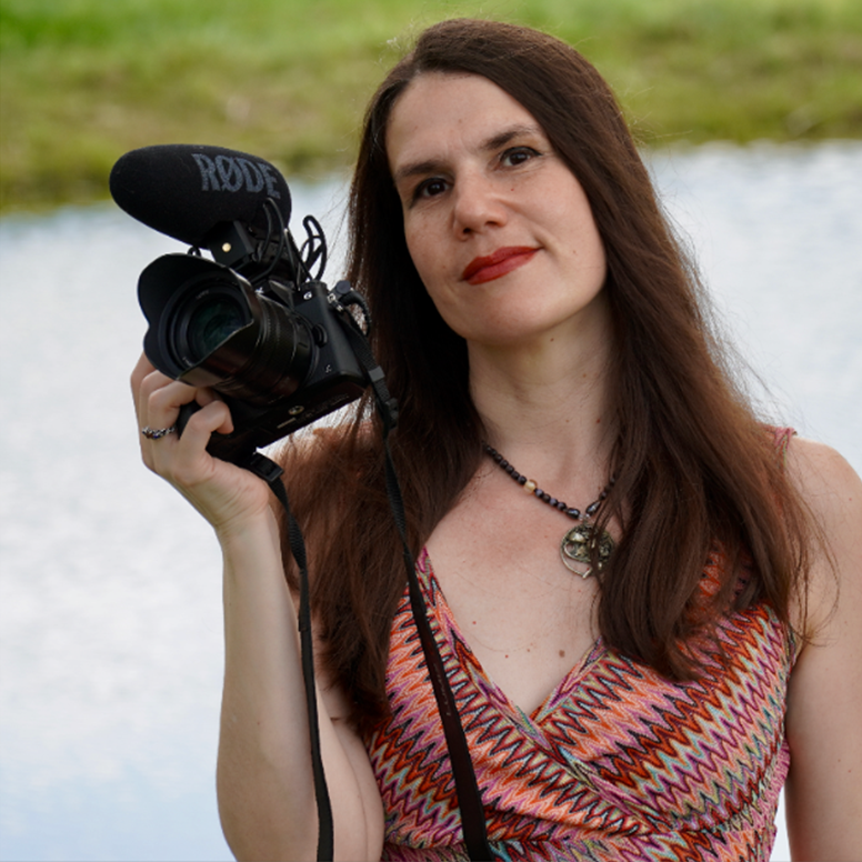Headshot of Alexandra Hidalgo. She is wearing a colorful, striped, short sleeve shirt and is holding up a camera with a mic on top.