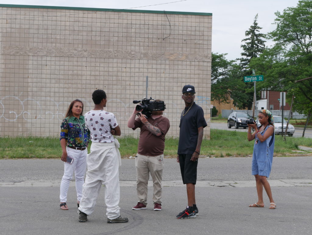 An image of people talking in a city parking lot. Geri is off to the left, listening intently while she talks with two men. There are two people filming the moment close up. 