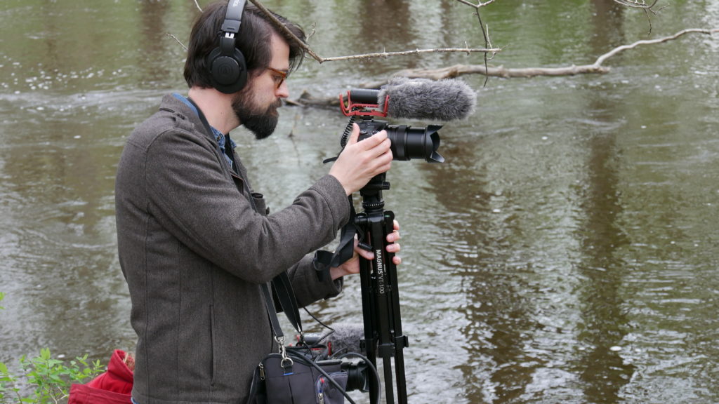 An image of Johnston angling a camera on a tripod. He is outside, standing by a river and wearing a gray-brown jacket. 