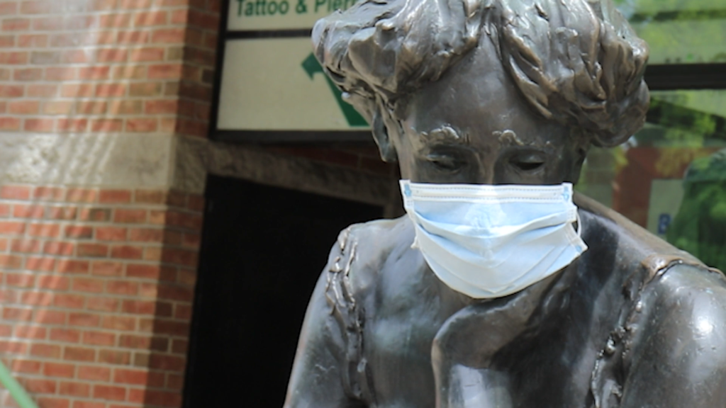 Image of a dark metal statue of a woman with a blue surgical mask on. 