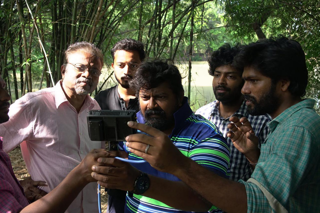 Swarnavel and others stand around a camera in what looks like a wooded area.