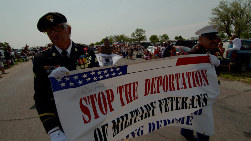 Still of two older veterans marching in uniform in a parade, holding a banner that says "Stop the deportation of military veterans and bring deported veterans home."